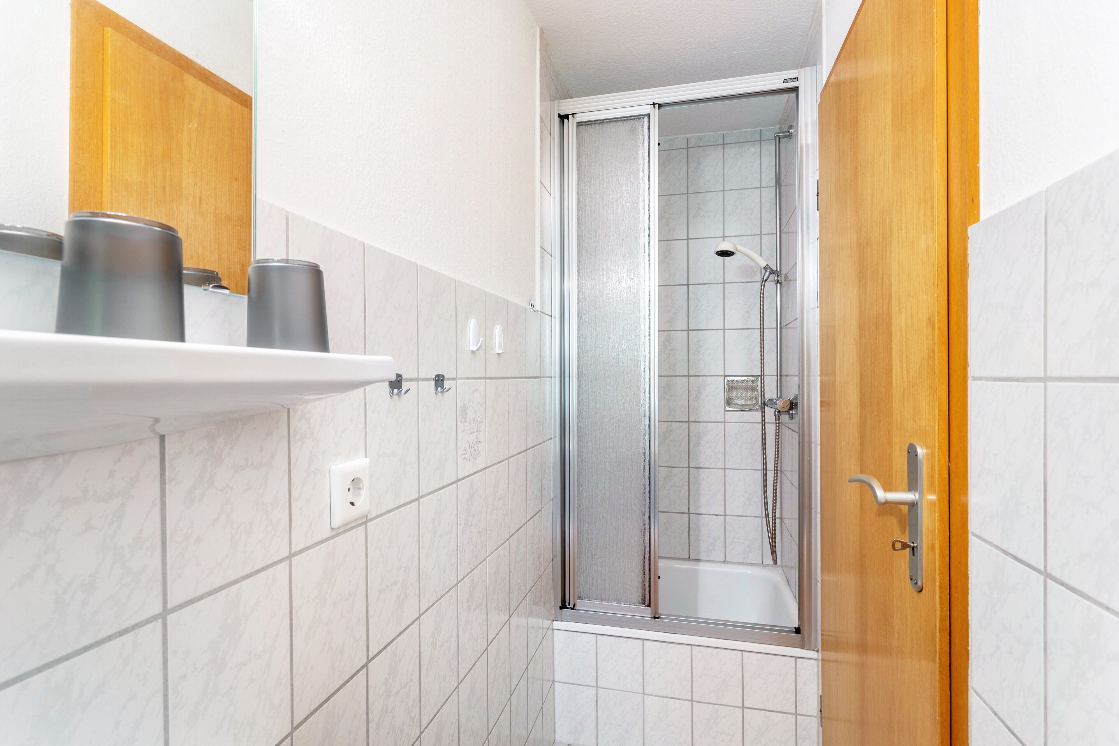 A bathroom with a shower, a wooden door, and a shelf with toiletries.