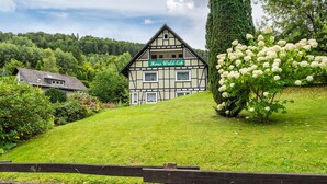 Une maison traditionnelle à colombages, sur une colline verdoyante bordée d’arbres, avec une pancarte indiquant « Haus Wald-Eck ». Une clôture en bois se trouve au premier plan.