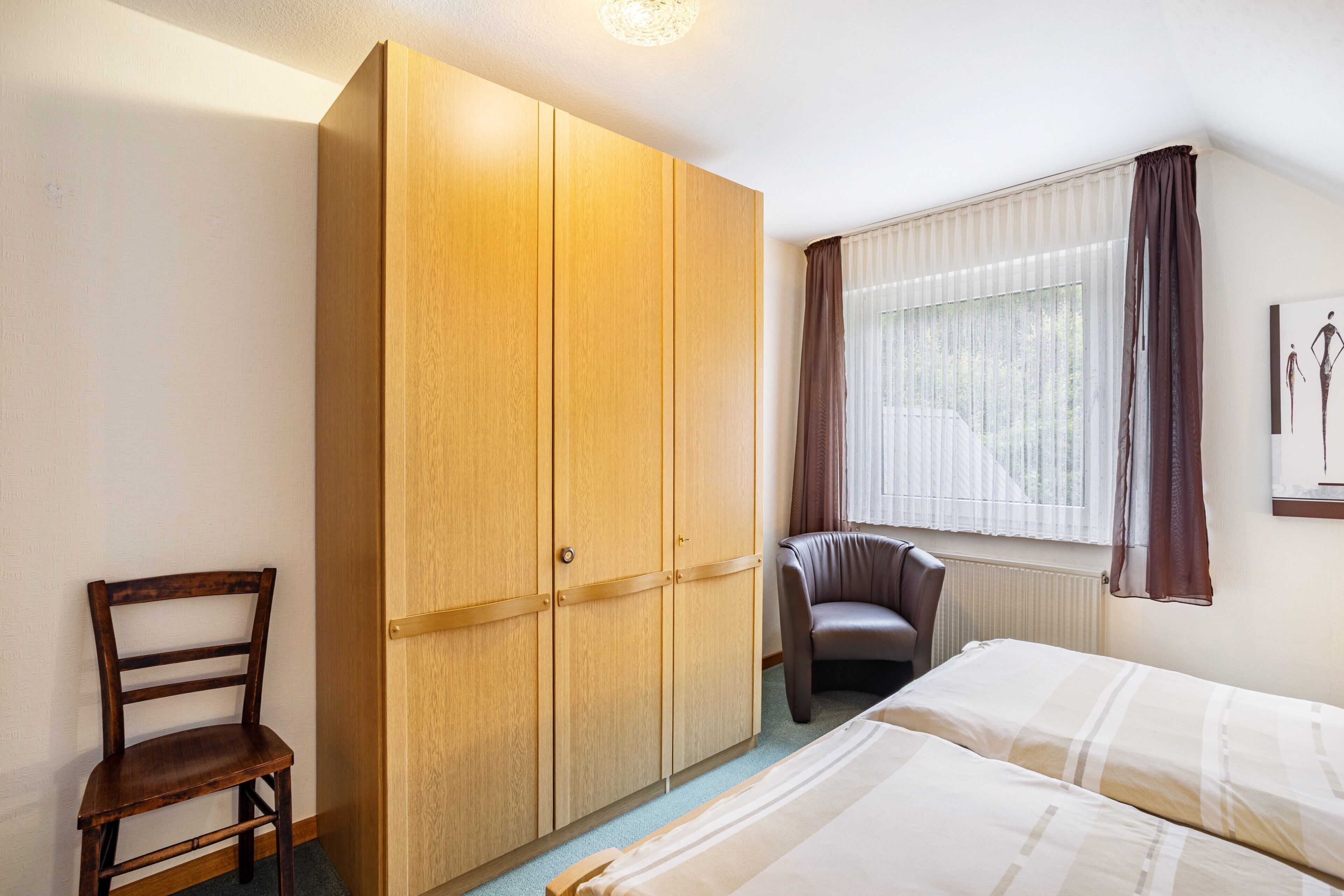 A bedroom with a bed, a wardrobe, a chair, and a window with curtains.