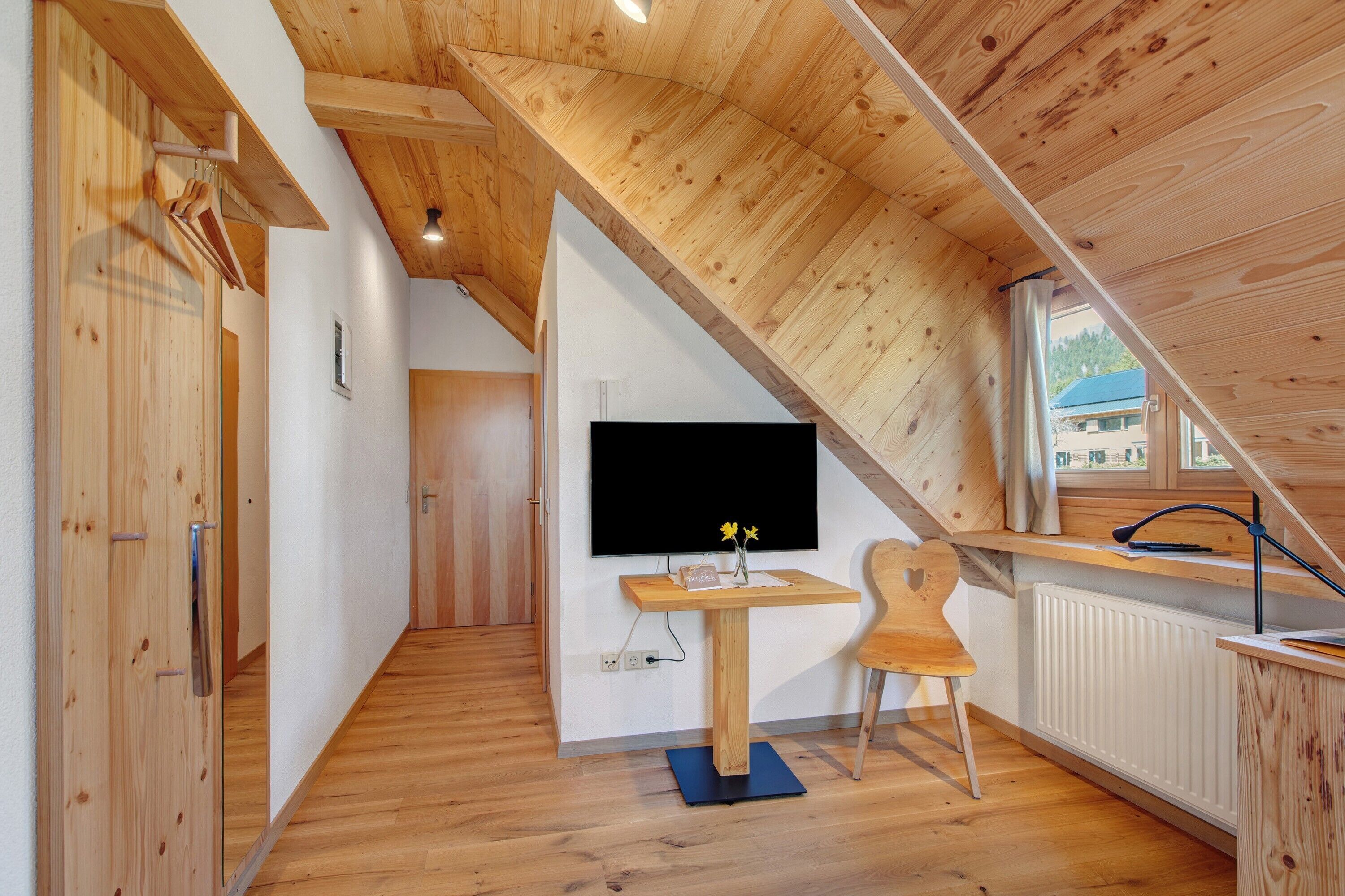 A wooden interior with a TV on a wall, a desk, a chair, and a radiator.