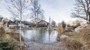 Un étang avec un pont en bois, entouré d’arbres et bordé d’un bâtiment doté d’un toit en pente.