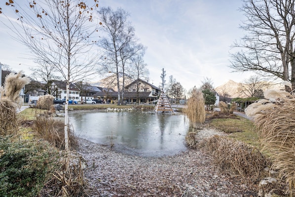 Un étang avec un pont en bois, entouré d’arbres et bordé d’un bâtiment doté d’un toit en pente.