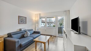 Un salon moderne avec un canapé, une table basse, un téléviseur et une fenêtre donnant sur des immeubles.