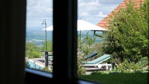 Blick durch ein Fenster auf einen Poolbereich mit Liegestühlen, eine überdachte Sitzgelegenheit und eine entfernte Landschaft.