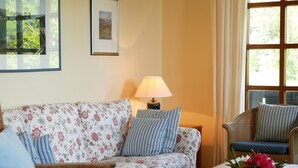 A living room with a floral sofa, a side table with a book, a lamp, and a window with curtains. - Apartment 'Bachblick' with Shared Pool and Wi-Fi (Sasbachwalden)