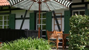A wooden bench under a striped umbrella, a ladder leaning against a wall, and a garden with green foliage. - Apartment 'Bachblick' with Shared Pool and Wi-Fi (Sasbachwalden)
