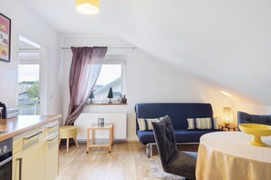 Una cocina moderna con encimera de madera, un taburete pequeño, una mesa de comedor con un centro de mesa amarillo, un sofá azul oscuro con almohadas a rayas, y una ventana con vistas a árboles y edificios.
