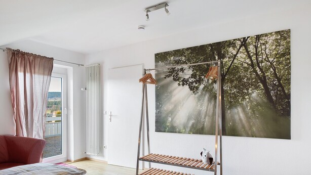 A room with a bed, a window with curtains, a wooden floor, a metal shelf, and a painting of trees. - Apartment 'Eulennestle' with Shared Terrace, Balcony and Wi-Fi (Lenzkirch)