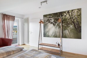 Una habitación con una cama, una ventana con cortinas, suelo de madera, una estantería metálica y un cuadro de árboles.