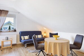 Een kamer met een bank, een ronde tafel, een klein tafeltje en een raam met uitzicht op een huis.
