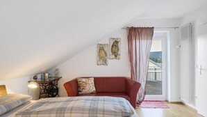 A bedroom with a bed, a red sofa, a nightstand with a lamp, and two wall-mounted owl paintings. - Apartment 'Eulennestle' with Shared Terrace, Balcony and Wi-Fi (Lenzkirch)