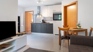 Eine moderne Küche mit grauen Schränken, einem Holzespeisetisch mit zwei Stühlen, einem Fernseher auf einem weißen Schrank und einer kleinen Pflanze auf dem Tisch.