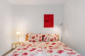 Une chambre à coucher avec un lit, une lampe de chevet, une œuvre d’art rouge accrochée au mur et une couvre-lit à motifs floraux.
