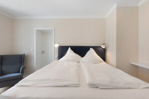Una cama doble con ropa blanca, un cabecero oscuro, lámparas de noche y un sillón gris.