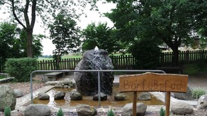 Uma placa de madeira com a inscrição 'Barfuf Bpfad' em ambiente natural, com uma pequena cascata, pedras e uma cerca de madeira ao fundo.