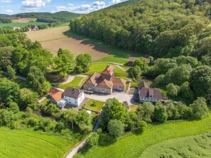 Vue aérienne d’un domaine rural comprenant plusieurs bâtiments, dont une grande maison en pierre et plusieurs constructions plus petites, le tout entouré d’une végétation luxuriante et de champs.