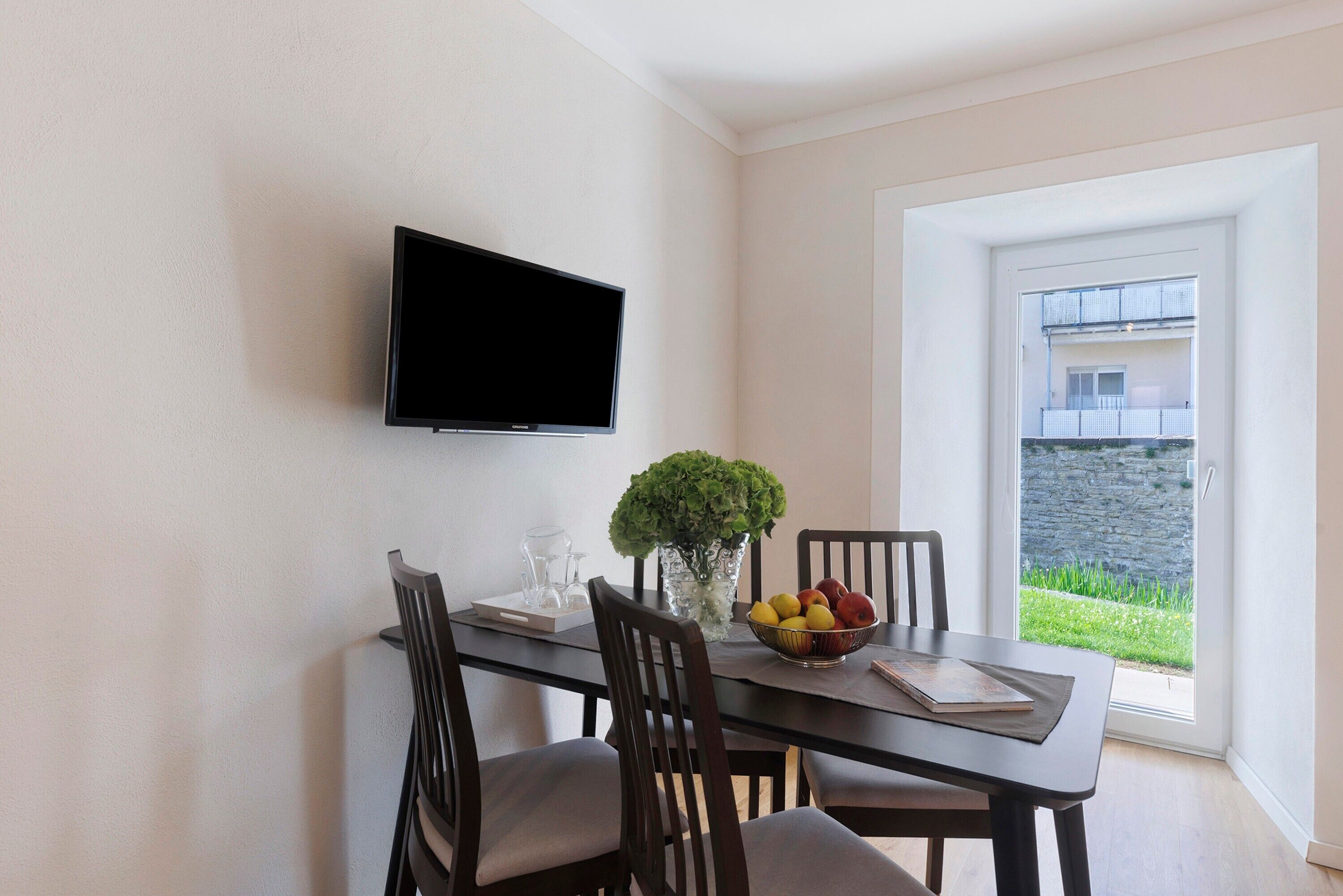 Een eetgedeelte met een tafel, stoelen, een flatscreen televisie aan de muur, een vaas met een plant en een schaal fruit.