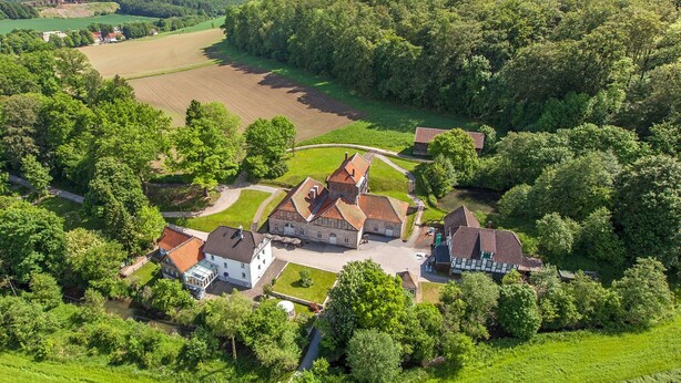 Vista aérea de una finca rural con varias construcciones, incluyendo una casa grande de piedra y otras estructuras más pequeñas, rodeada de abundante vegetación y campos.