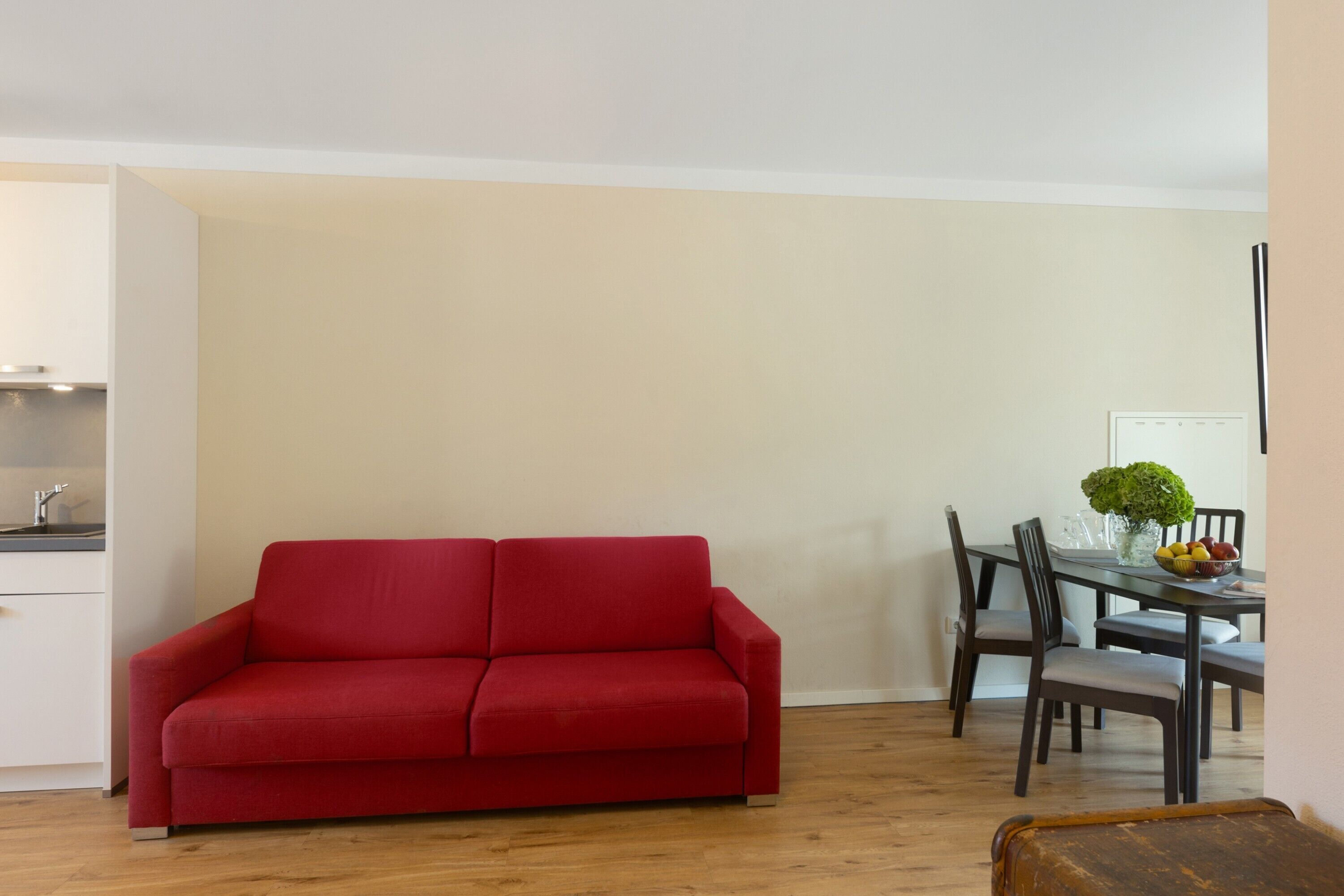 A red sofa in a room with a dining table and chairs, a vase with a plant, and a wooden floor.