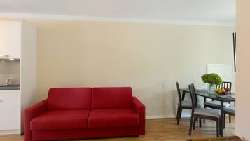 A red sofa in a room with a dining table and chairs, a vase with a plant, and a wooden floor.