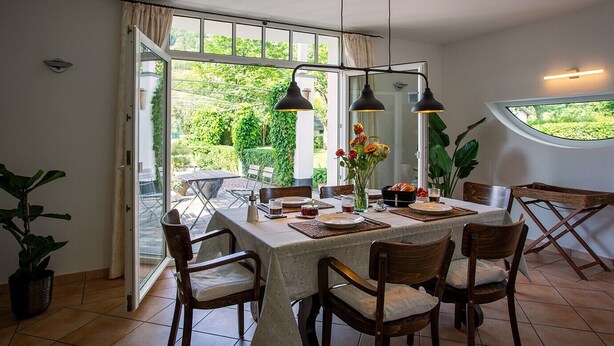 A dining area with a table set for four, two pendant lights, a vase with flowers, and a view of greenery through large windows. - Holiday Home 'Wennetraum' with Sauna, Terrace, and Garden (All for Private Use!) (Eslohe)