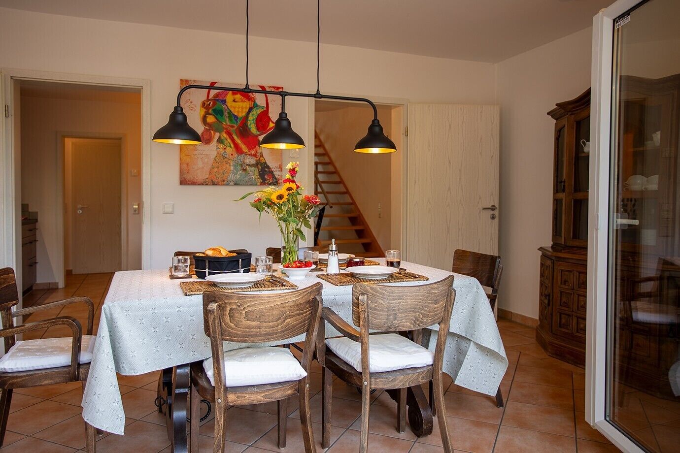 Un comedor con una mesa preparada para cuatro, sillas, un cuadro en la pared y una escalera al fondo.