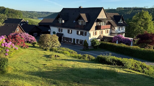A large, two-story house with a steeply pitched roof, surrounded by lush greenery and flowering plants. - Apartment '4, Größe 60qm' with Balcony and Wi-Fi (Herscheid)