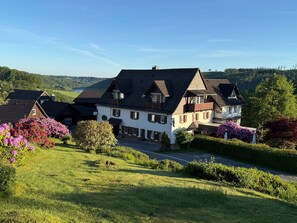 Una grande casa a due piani con un tetto spiovente, circondata da un lussureggiante verde e piante fiorite.