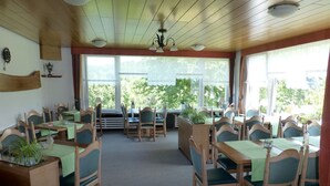 A dining area with wooden tables and chairs, large windows with a view of greenery, and ceiling fans. - Apartment '4, Größe 60qm' with Balcony and Wi-Fi (Herscheid)