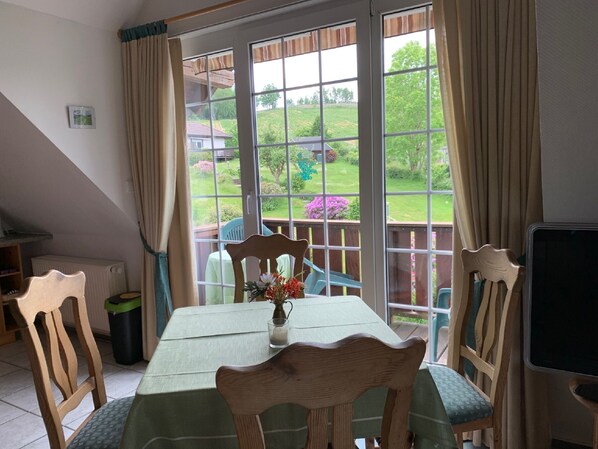 A dining area with a table and chairs, a view of a garden, and a window with curtains. - Apartment '4, Größe 60qm' with Balcony and Wi-Fi (Herscheid)