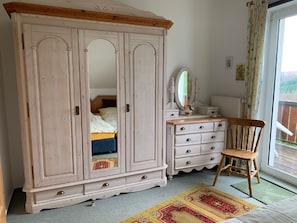 Ein Schlafzimmer mit großem Kleiderschrank, Nachttisch mit Spiegel, einem Sessel und Blick ins Freie durch ein Fenster.