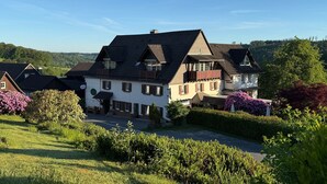 Een groot, ruim tweelaags huis met een schuin dak, omgeven door weelderige begroeiing en een heldere lucht.