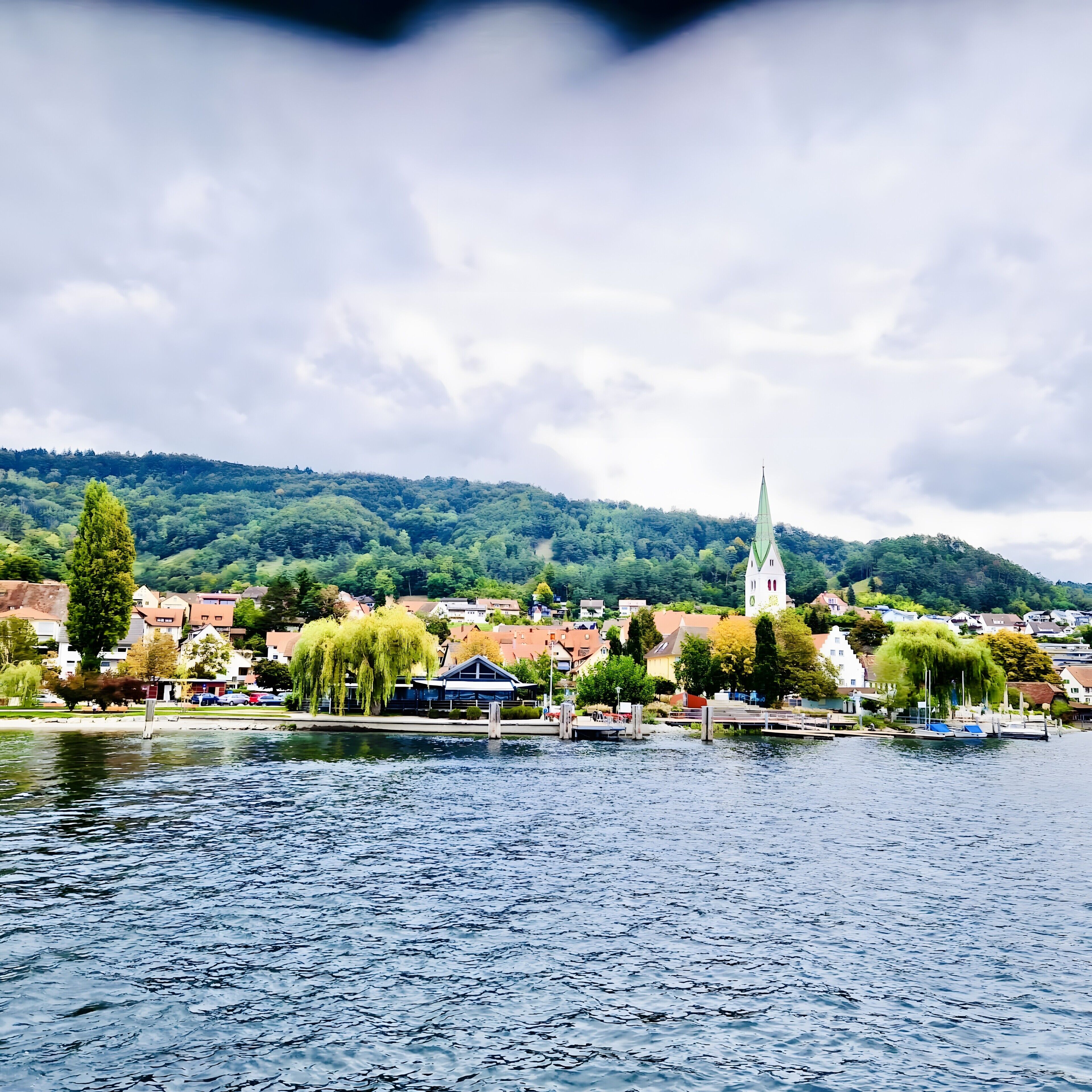 A lakeside town with a prominent church steeple, calm water, and surrounding greenery.