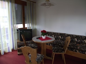 A room with a television, a round table with chairs, a sofa, and a window with blinds.