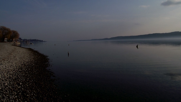 Un lac paisible, bordé d’une plage de galets, avec des collines qui se dessinent à l’horizon.