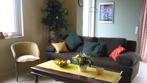 A living room with a sofa, a chair, a coffee table, a plant, and a bowl of fruit.