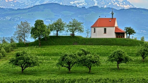 A single-story building with a red roof, surrounded by green fields and trees, with a mountain range in the background. - Apartment 'Stapelfeldt' with Private Terrace, Shared Garden and Wi-Fi (Wasserburg (Bodensee))