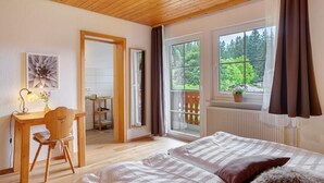 Un dormitorio con techo de madera, cama con ropa de cama estampada, escritorio con silla y vista a la vegetación a través de grandes ventanales.