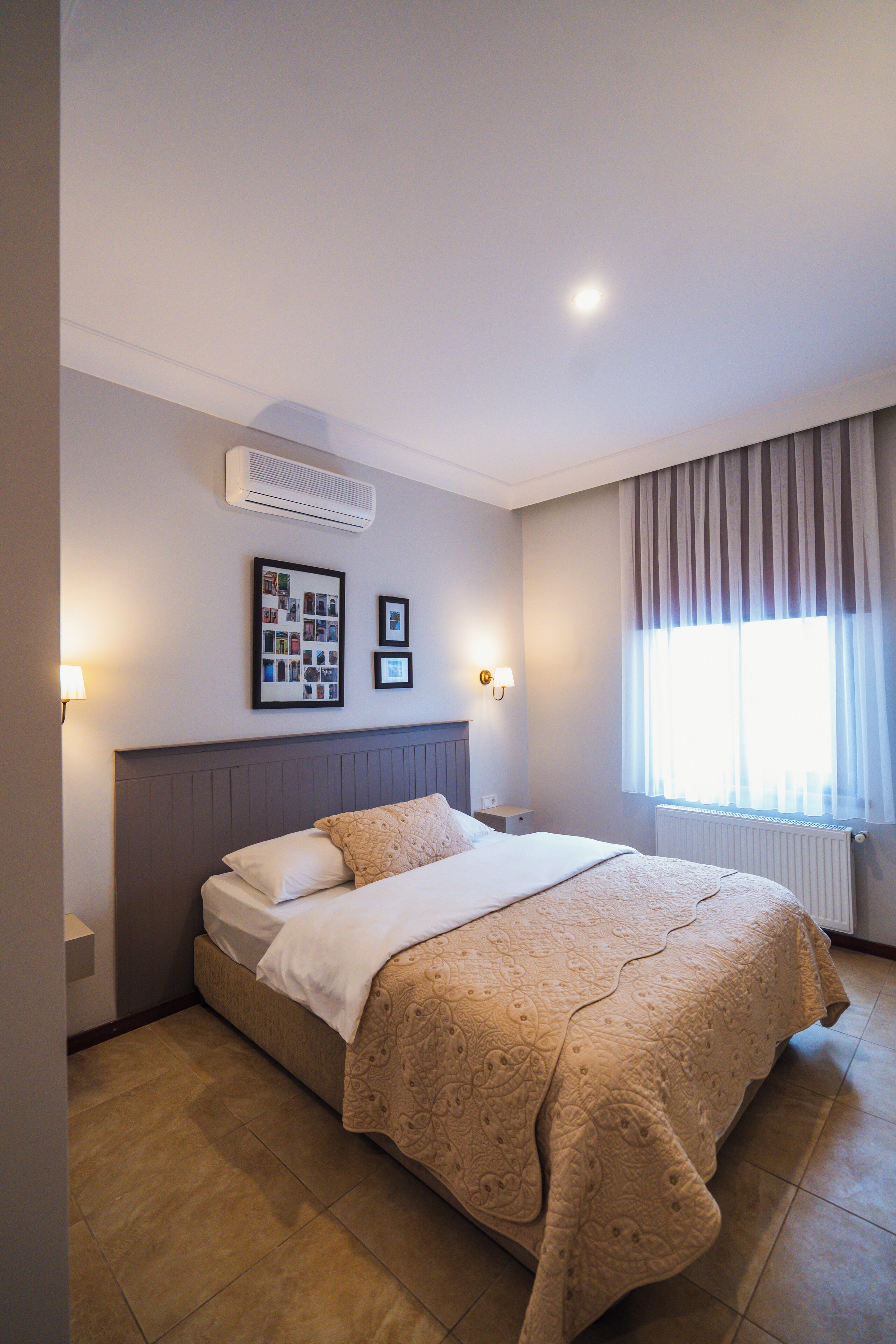 A neatly made bed with a patterned bedspread, a wall-mounted air conditioner, and two framed pictures above it.