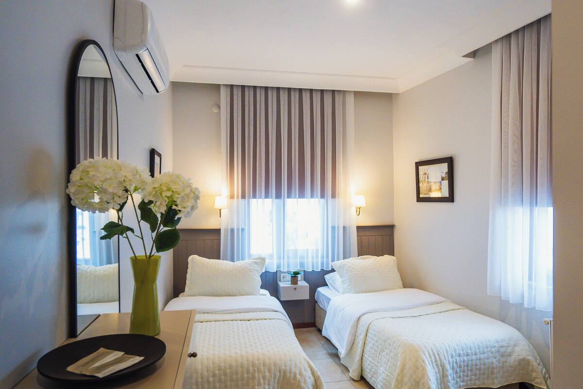 A hotel room with two beds, a desk, a mirror, a vase with flowers, and a window with curtains.