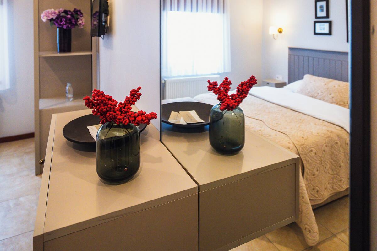 A modern hotel room with a bed, bedside tables, a mirror, and a vase with red flowers.