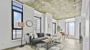 A modern living room with a grey sofa, a coffee table, and a rug. There are large windows, a dining area, and a kitchen visible.