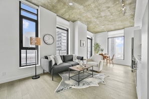A modern living room with a grey sofa, a coffee table, and a rug. There are large windows, a dining area, and a kitchen visible. - Calgary Tower View | King Bed | Mins to Saddledome (Calgary)