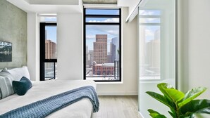 A modern bedroom with a large bed, a view of the city through a window, and a potted plant.
