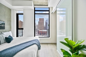 A modern bedroom with a large bed, a view of the city through a window, and a potted plant. - Calgary Tower View | King Bed | Mins to Saddledome (Calgary)