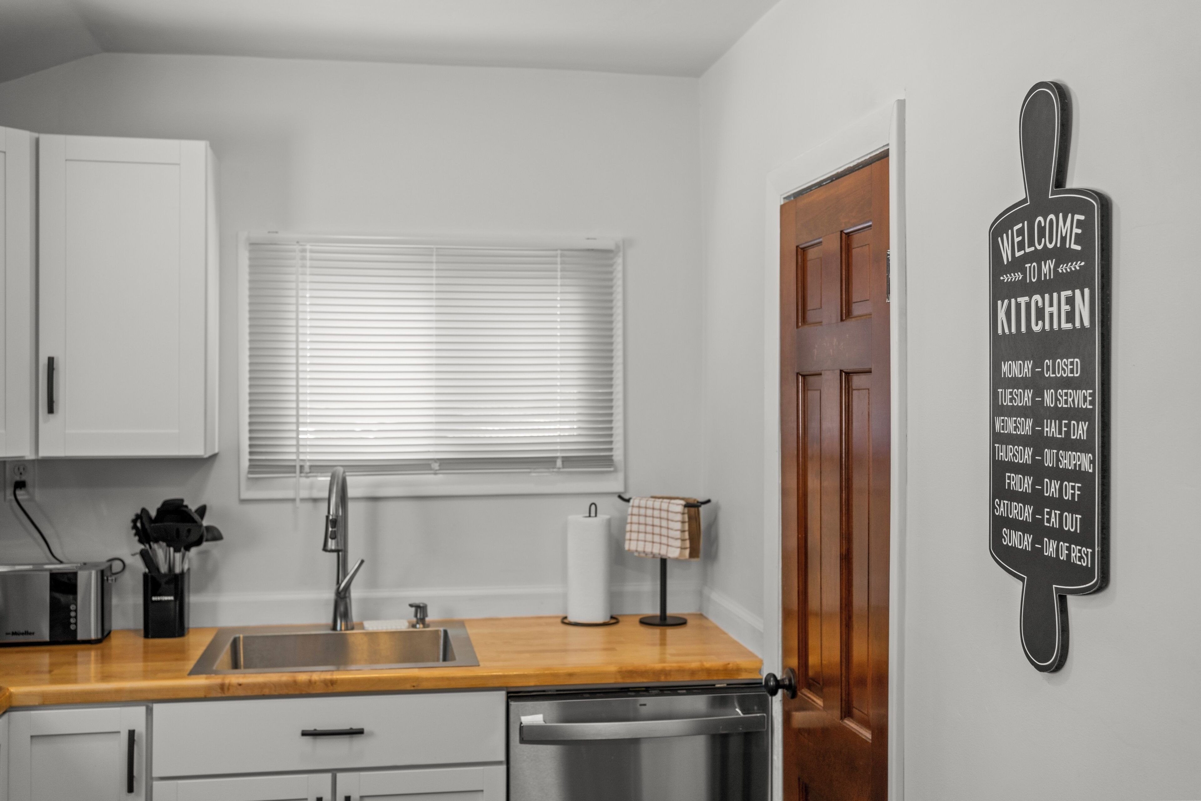 A kitchen with a wooden countertop, a sink, a dishwasher, and a window with blinds. There is a sign on the wall that says "Welcome to my Kitchen" with a schedule of days and activities.