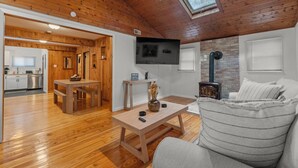 A cozy living room with a wooden ceiling, a TV mounted on the wall, a fireplace, and a dining area with a table and bench.