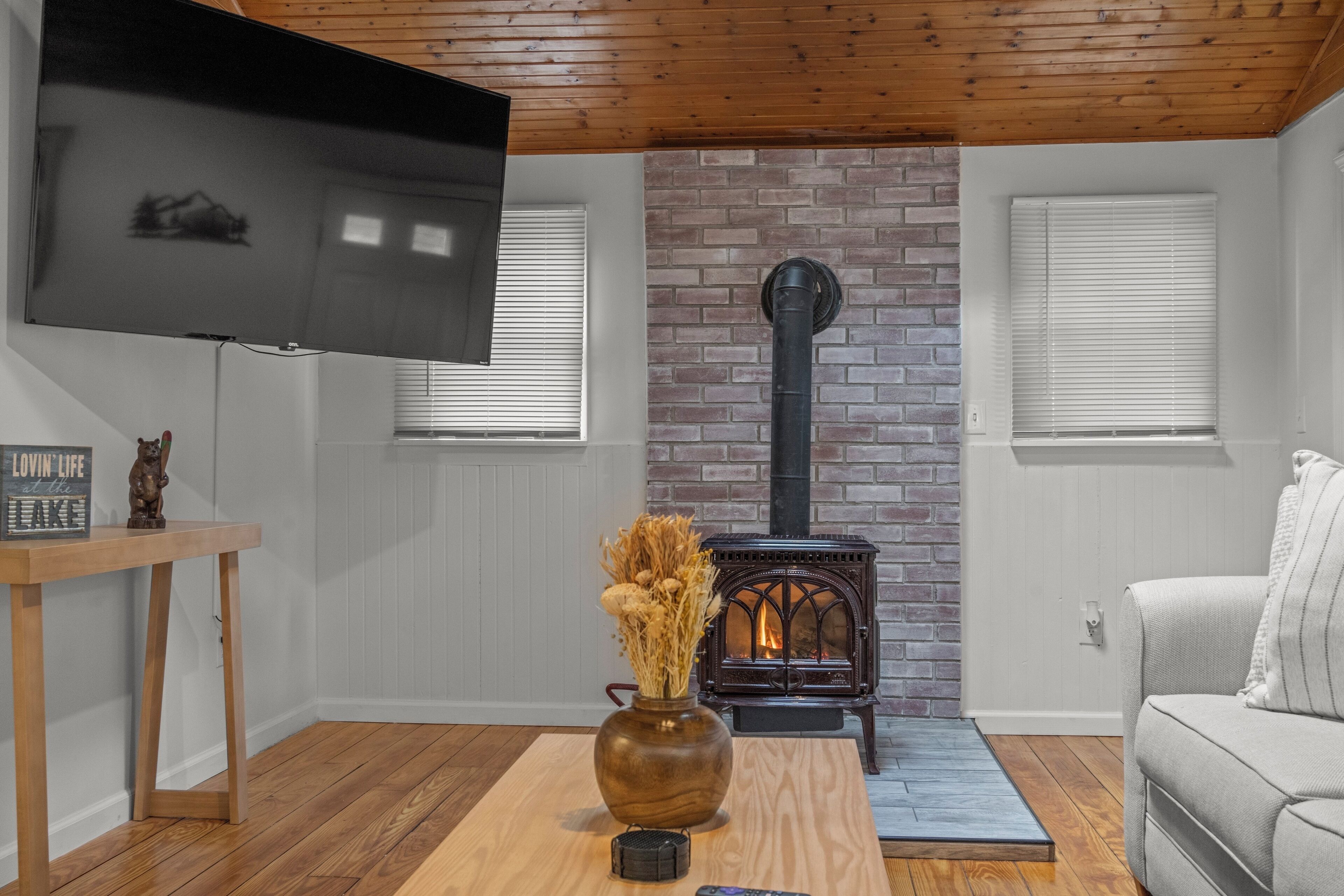 A living room with a brick fireplace, a flat-screen TV mounted on the wall, a wooden coffee table with a vase of dried flowers, and a light-colored sofa.