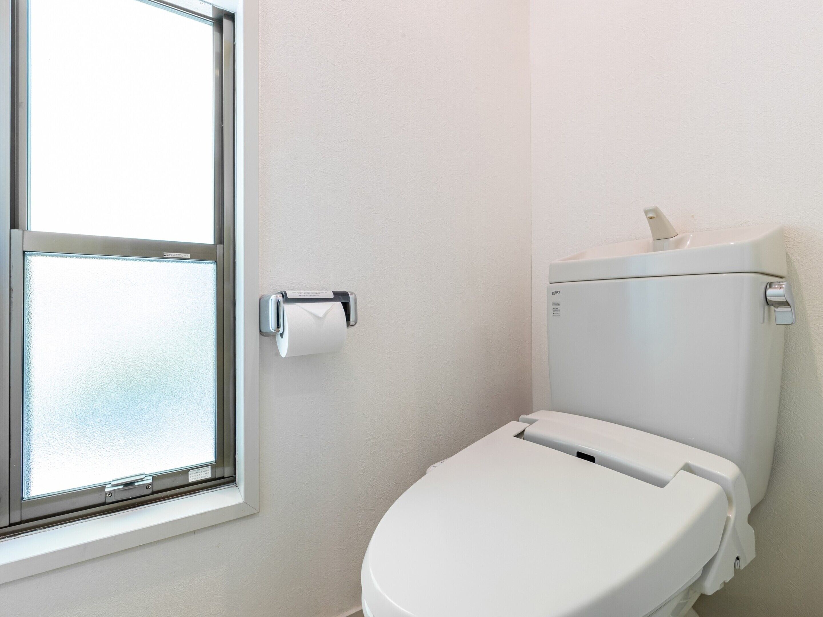 A white toilet with a paper towel dispenser, a window with a view of the outside, and a white wall.