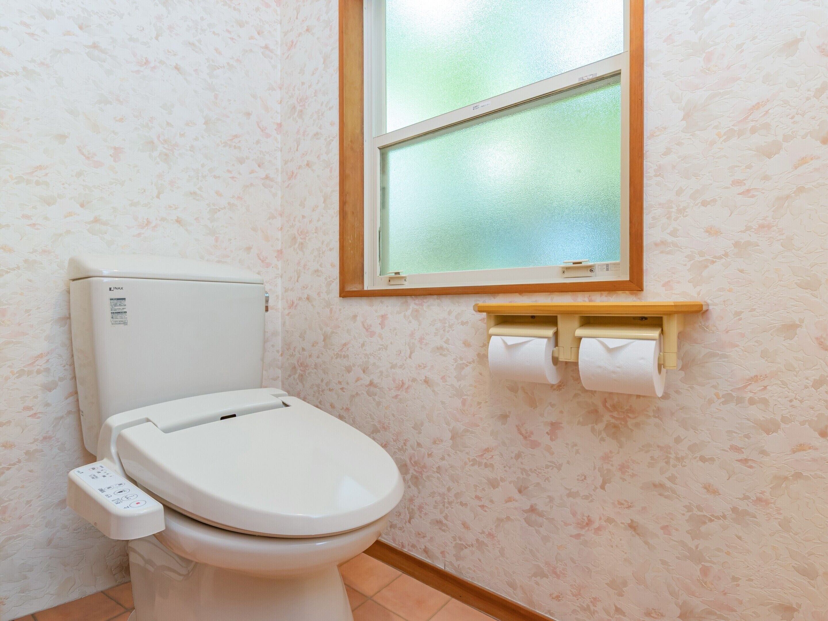 Sanitário com painel de controle, uma janela acima e rolo de papel higiênico na parede.